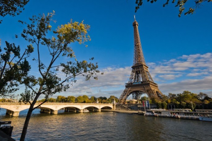 Eiffel Tower with trees and river in foreground under a blue sky.