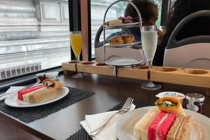 Tea set with sandwiches and pastries on a table inside a bus with a city view.