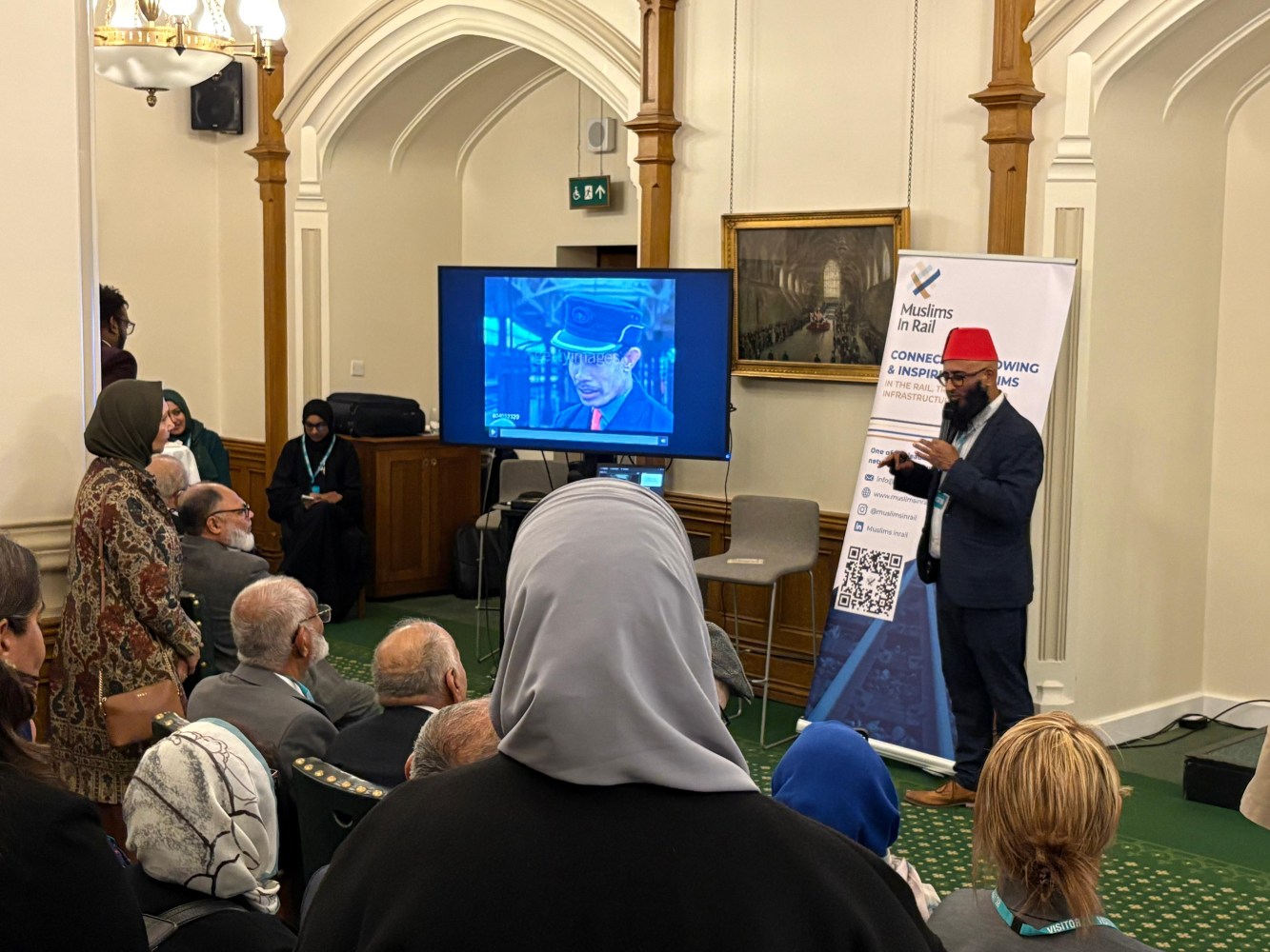 A speaker presents to an audience in a room with a banner reading 'Muslims in Rail' and a screen showing a video.