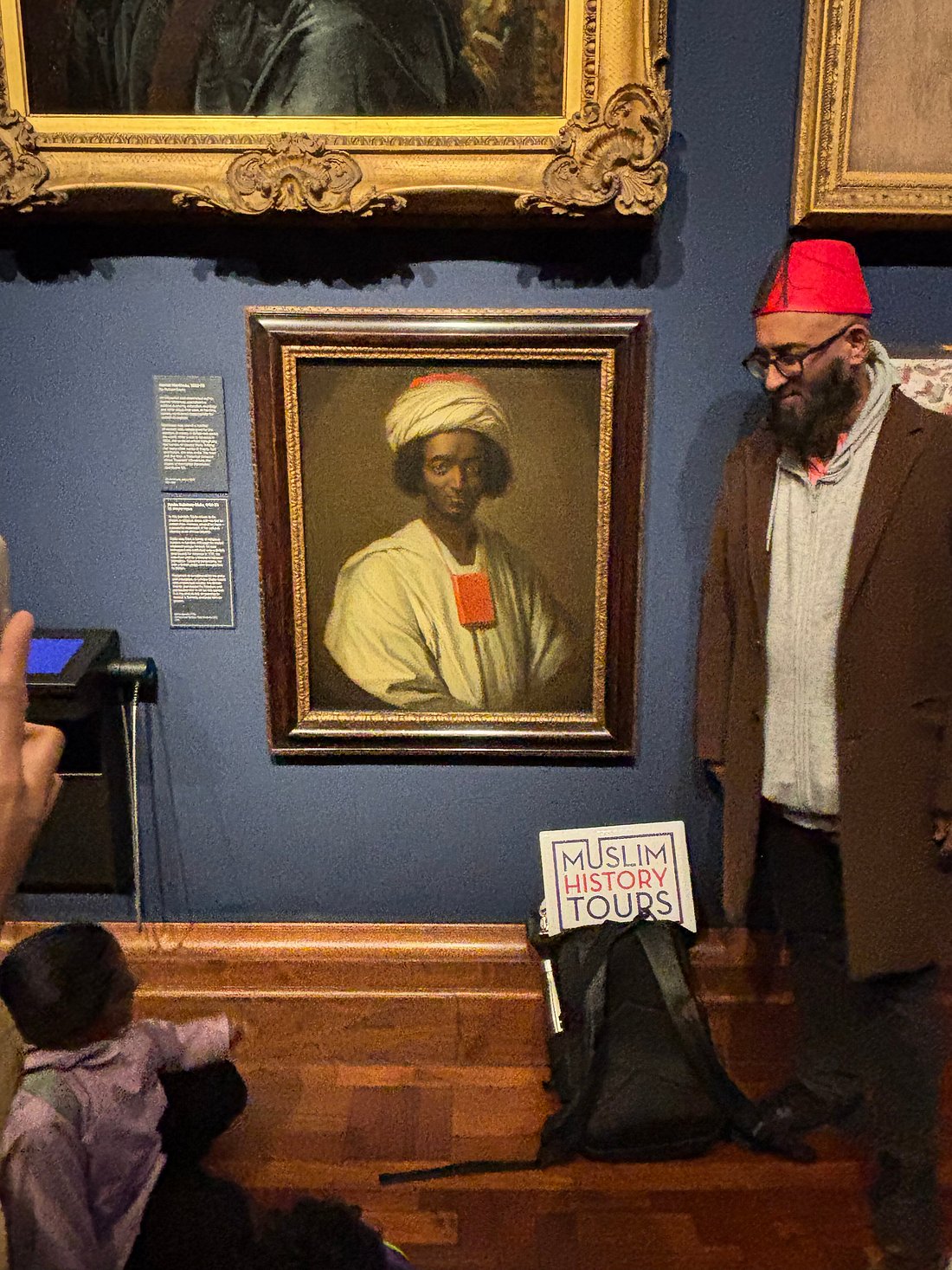 Man in red hat next to portrait in museum, with sign reading 'Muslim History Tours' on a bag.