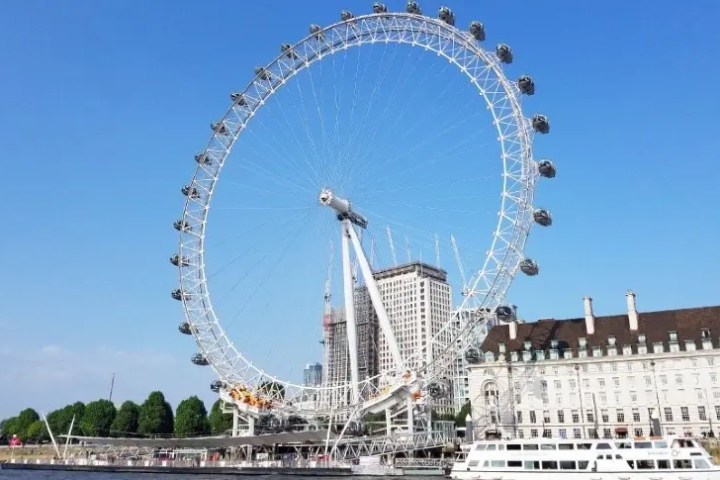 Large Ferris wheel by river with buildings and trees under a clear blue sky.