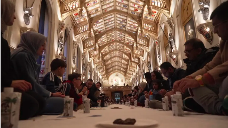 People sit on both sides of a long table in a grand hall with ornate ceilings and stained glass.