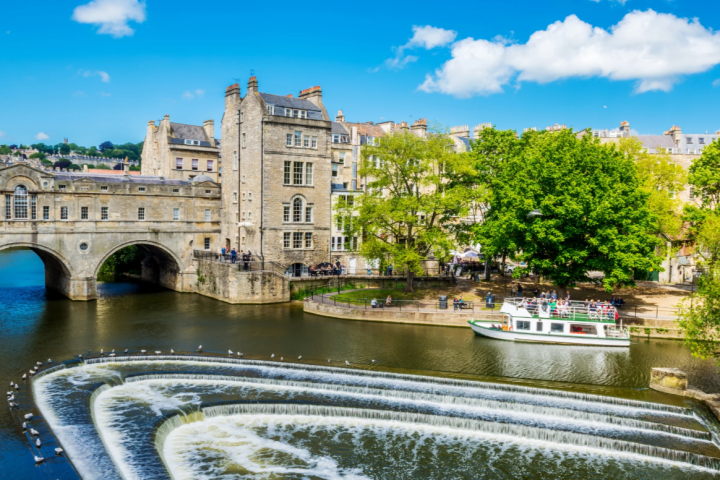 Scenic river view with bridge, buildings, trees, and a tour boat under a blue sky.