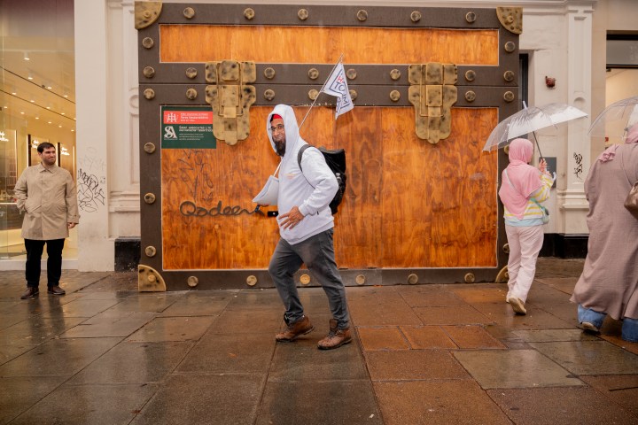Man in white hoodie poses in front of a wooden facade; people with umbrellas walk by.