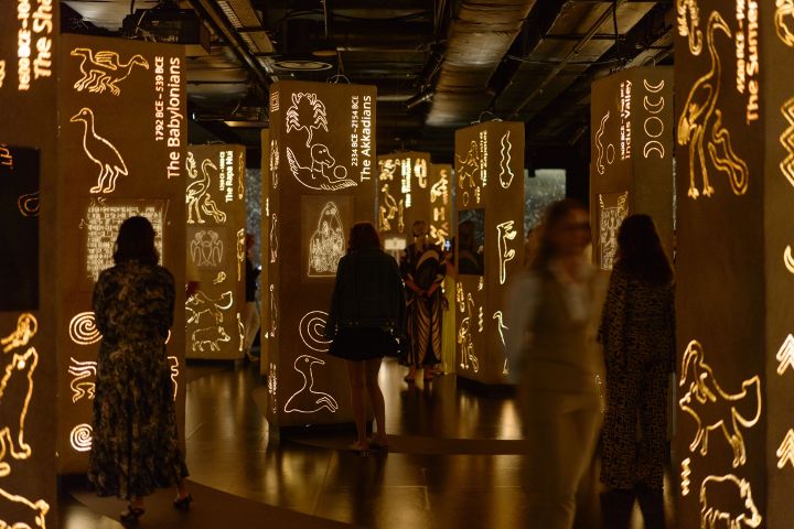 People in dimly lit exhibition with glowing columns displaying ancient symbols and figures.