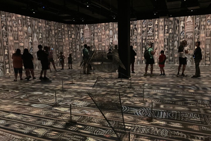 People in a dim room with patterned walls and floor, observing an art installation.
