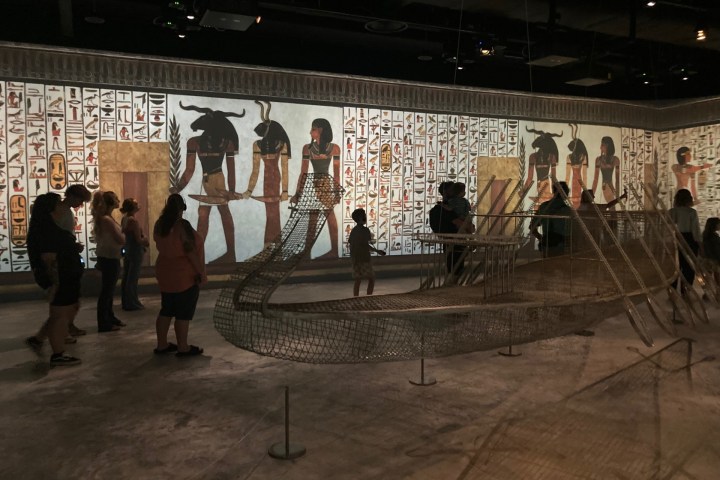 People view wire sculptures and Egyptian mural in a museum exhibit.