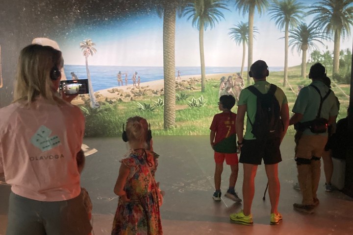 People watching a tropical beach scene on a large screen with palm trees and ocean.