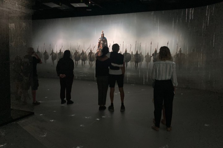 People viewing a lit scene of warriors with shields in a dark room.