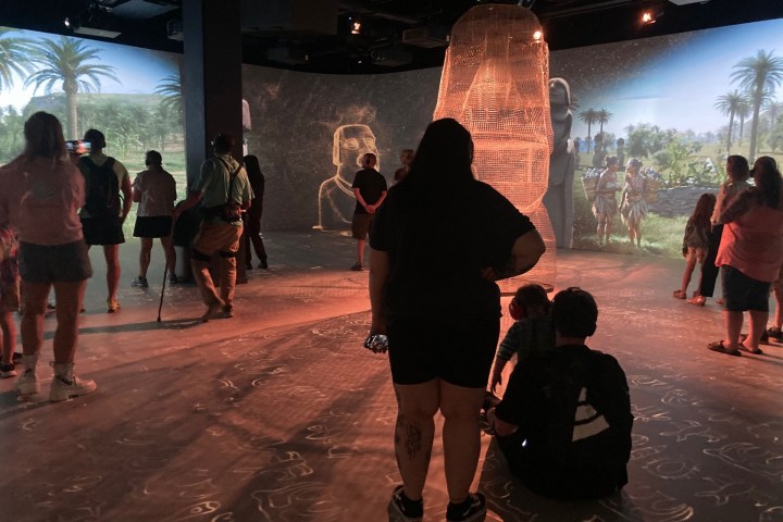 People viewing an immersive digital display of ancient sculptures and landscapes.