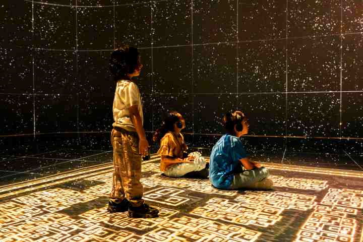 Three children with headphones watch a starry projection in a dark room.