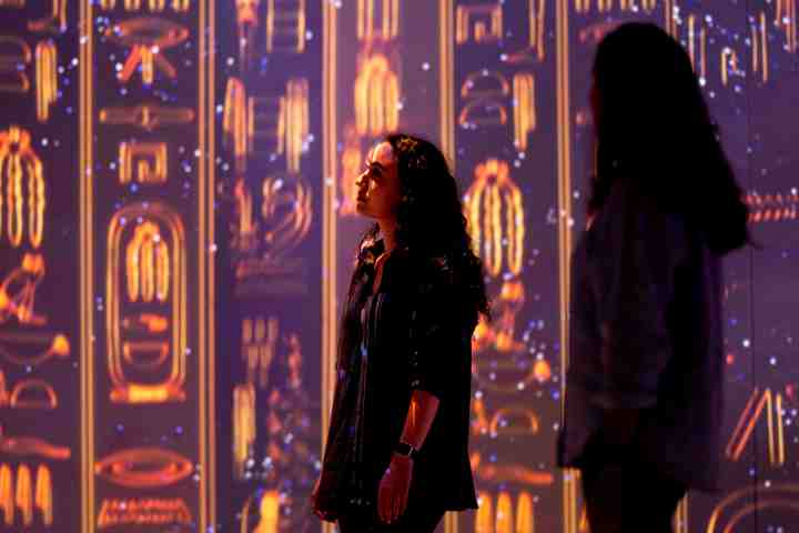 Two people stand in a room with illuminated Egyptian hieroglyphics on walls.