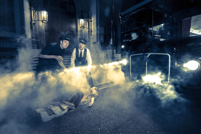 Two men in vintage attire investigate a lifeless figure on street, surrounded by smoke and a classic car.