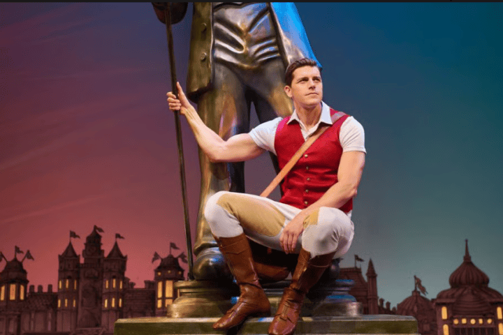 A man in period costume poses seated near a large statue, with a night cityscape in the background.